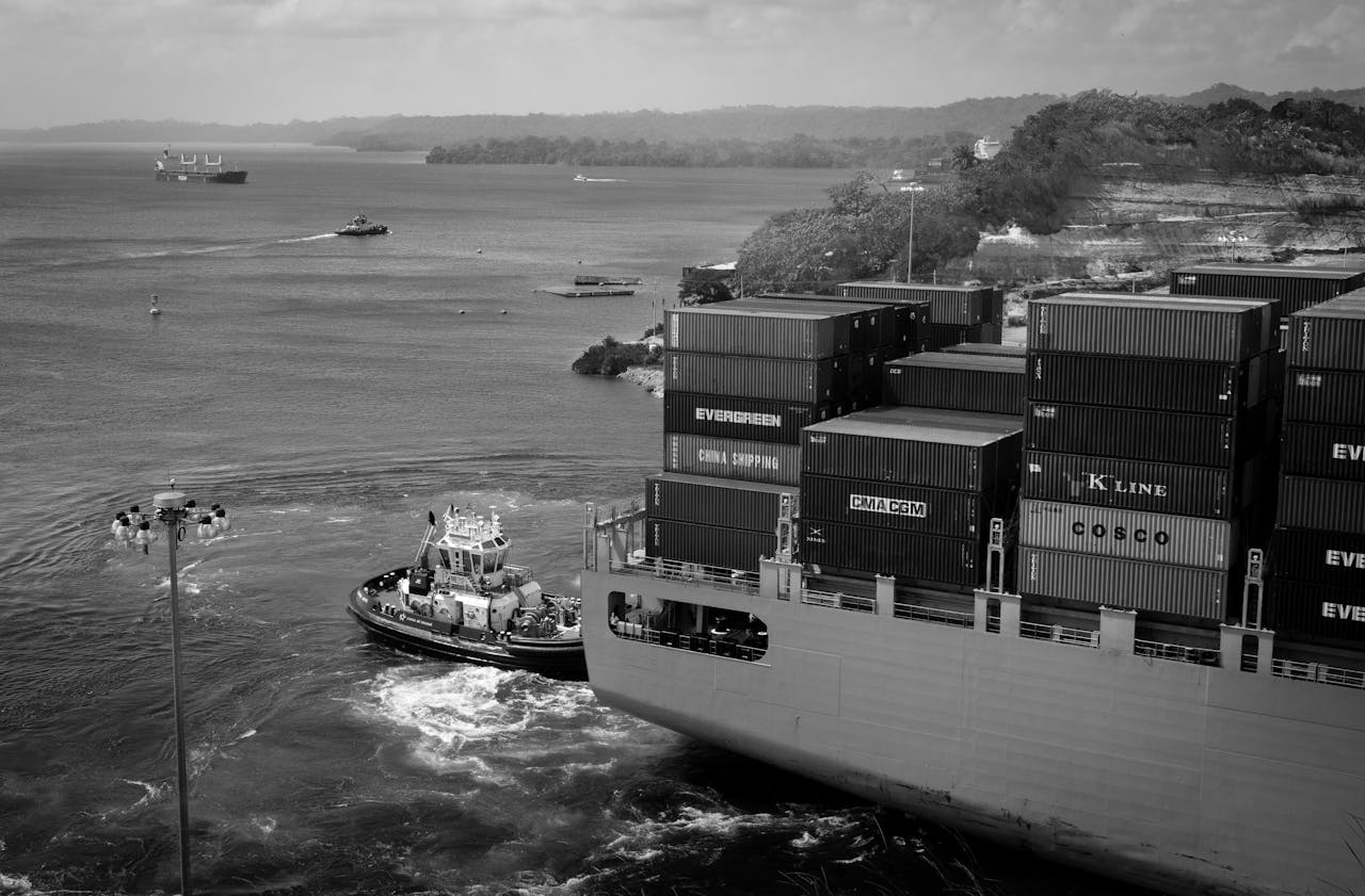 services-05 Black and white contemporary cargo vessel with many containers floating on calm river near hilly grassy shore towards industrial port